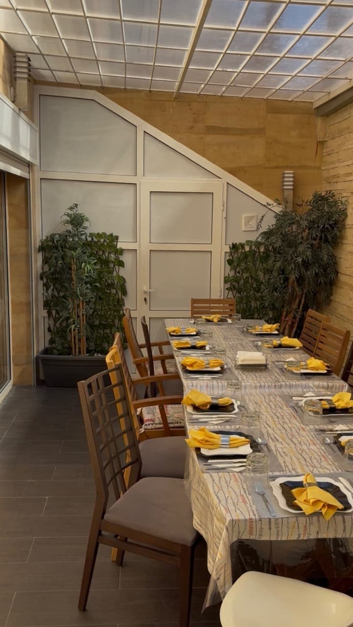 Longue table dressée avec serviettes jaunes, chaises en bois et plantes dans une véranda lumineuse.