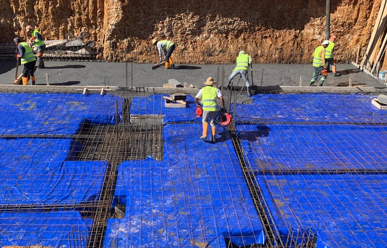 Ouvriers en gilets jaunes coulant du béton sur une fondation recouverte de bâches bleues.