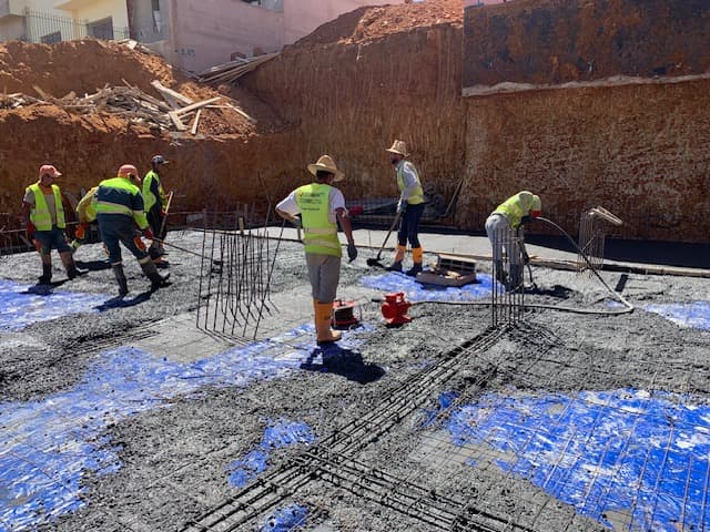Ouvriers en gilets jaunes coulant du béton sur une armature métallique et bâche bleue.