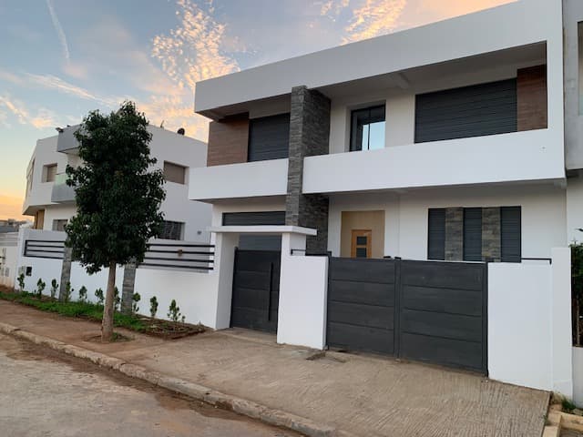 Maison blanche moderne avec détails en pierre, portail noir et petit arbre au crépuscule.